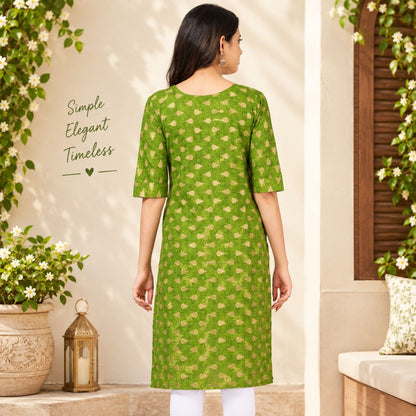 A woman stands with her back to the camera wearing a green knee-length kurta with a gold floral pattern, set against a warm beige wall decorated with green foliage and a lantern.