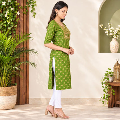 A woman modeling a green printed kurta with gold embroidery on the neckline, paired with white pants and beige heeled sandals, standing in a styled indoor setting with plants and decorative elements.