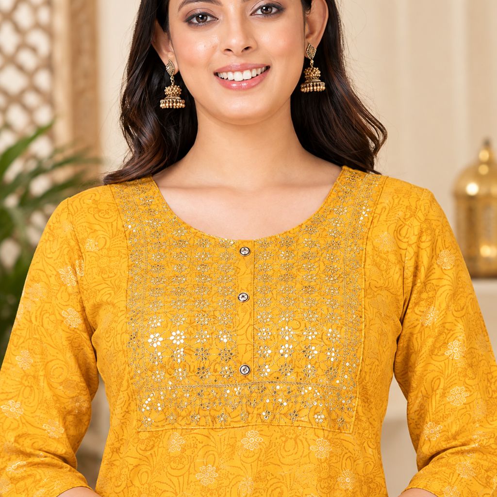 A woman wearing a mustard yellow embroidered kurta with silver mirror work and three buttons, paired with gold jhumka earrings, in a warm indoor setting.