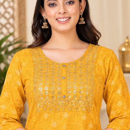 A woman wearing a mustard yellow embroidered kurta with silver mirror work and three buttons, paired with gold jhumka earrings, in a warm indoor setting.