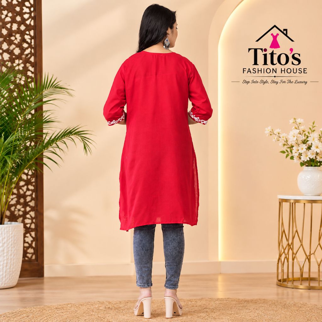 A woman is shown from the back wearing a bright red tunic with three-quarter sleeves and lace-trimmed cuffs, paired with blue jeans and white heels. The setting is a warm, modern indoor studio with beige walls, a potted plant, and a side table with flowers.