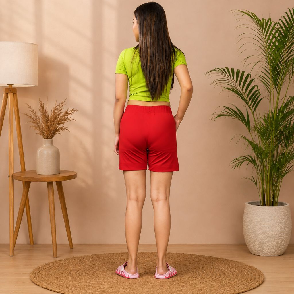 A woman stands with her back to the camera wearing a lime green short-sleeve top and bright red athletic shorts in a styled indoor room with a lamp, side table, dried plant, and potted palm.