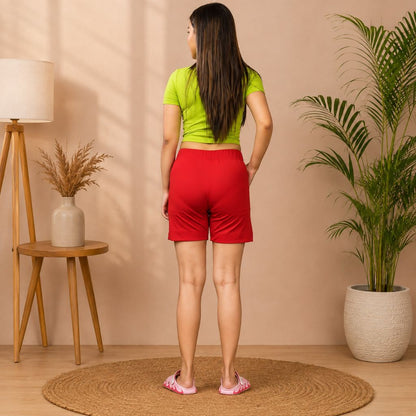 A woman stands with her back to the camera wearing a lime green short-sleeve top and bright red athletic shorts in a styled indoor room with a lamp, side table, dried plant, and potted palm.