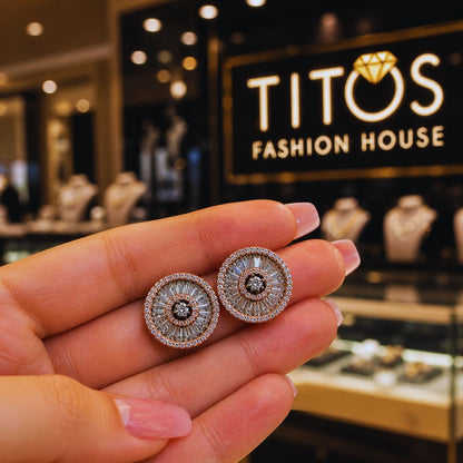 Close-up of a hand holding a pair of diamond earrings with 'TITOS Fashion House' in the background.